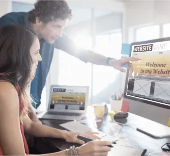 Two web designers collaborate on a 'WEBSITE GAME' homepage design displayed on a computer screen.