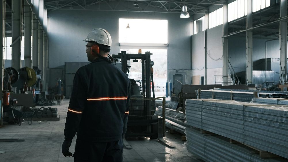 Worker in hard hat walks through a dimly lit industrial warehouse with machinery.
