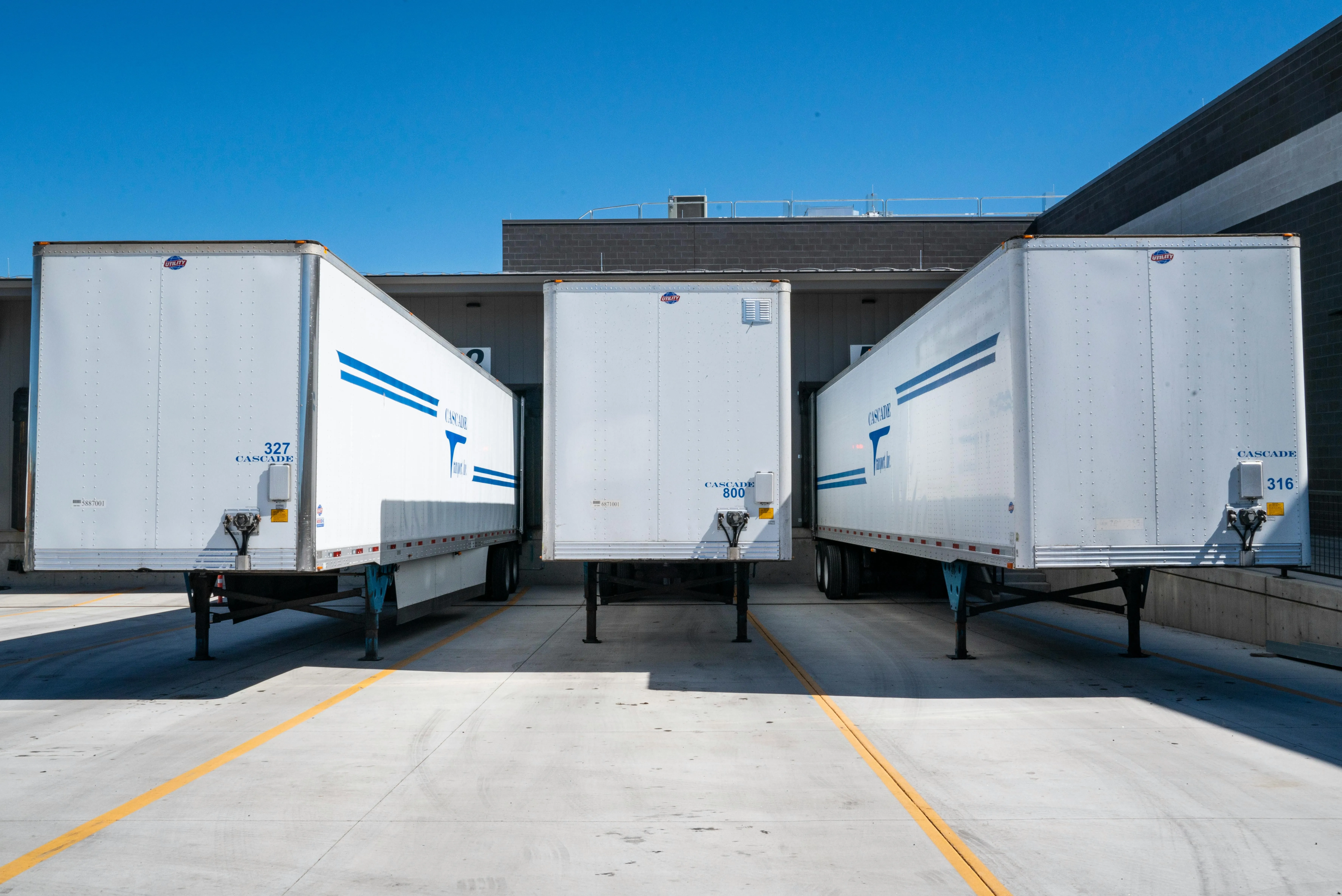 Three white semi-trailers backed into a loading dock under a bright blue sky.