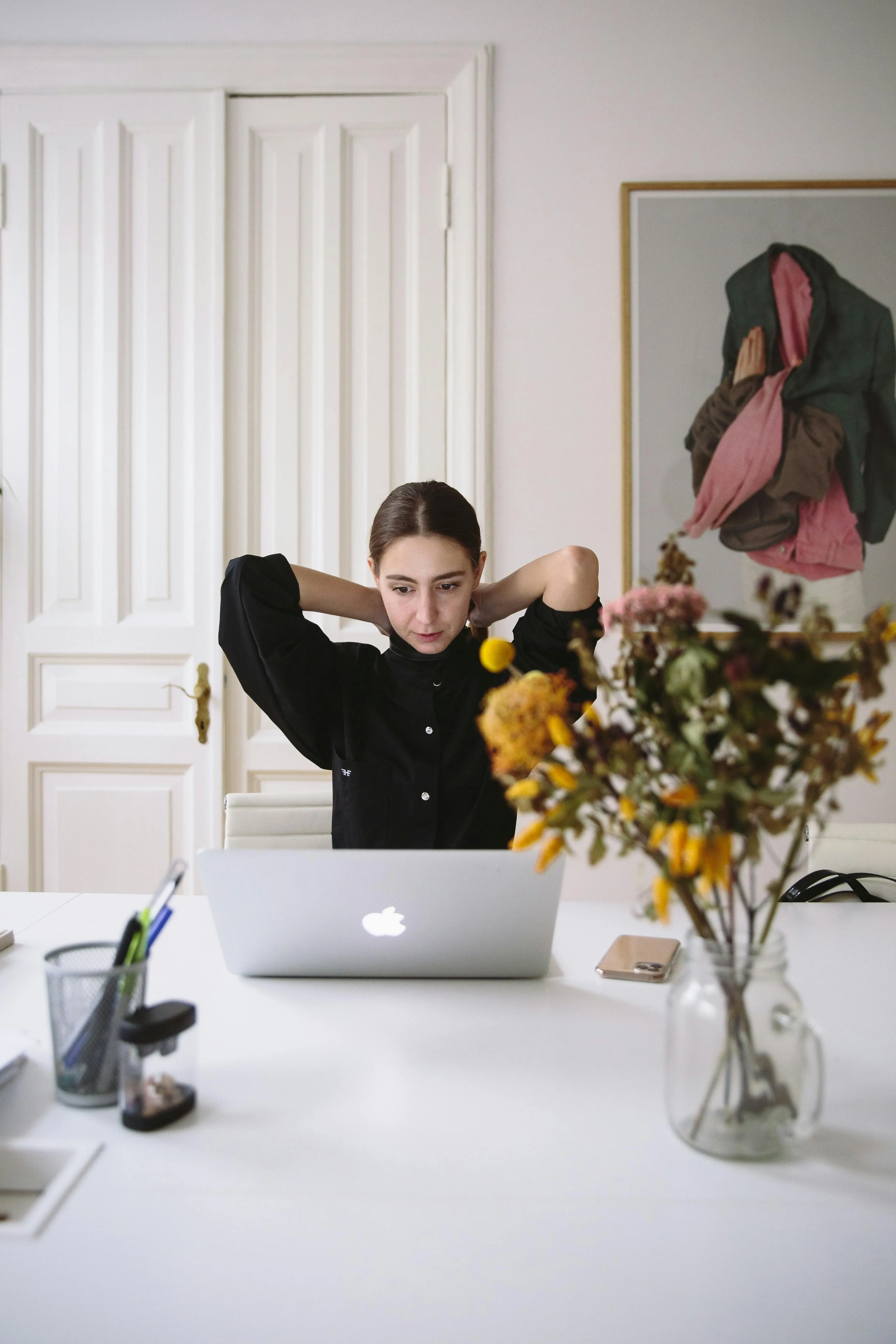 Young woman stretching with hands behind head at laptop on a white desk with flowers.