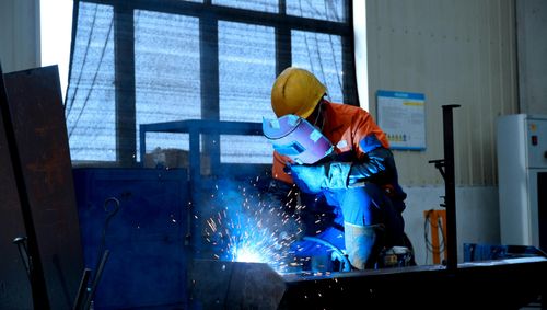 Worker in protective gear welds metal, creating bright sparks in a workshop environment.